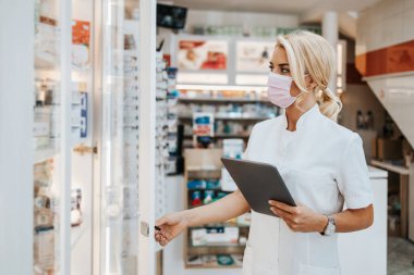 Young and attractive female pharmacist with face protective mask working in drugstore. She is confident and serious. Covid-19 open for business concept. clipart