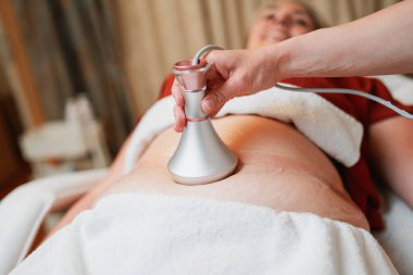 Güzellik uzmanı, profesyonel güzellik merkezinde yatan gülümseyen olgun bir kadının karnına radyasyon kaldırma cihazı uyguluyor. Modern, zararsız bir vücut şekillendirme tedavisi sunuyor..
