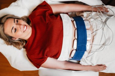 Rahatlamış bir kadın spa yatağında yatıyor, karnında lipolizis tedavisi görüyor, cildine birden fazla aplikatör takılmış, mavi kemer ile.