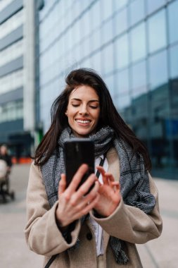 Şehir caddesinde dikilen güzel modaya uygun bayan turist çevrimiçi iletişim ve sosyal medya içeriği oluşturmak için elektronik akıllı telefon kullanıyor..