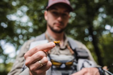 Angler focuses on showcasing a brightly colored artificial fly lure held between his fingers, highlighting preparation and detail before fly fishing in a natural outdoor setting