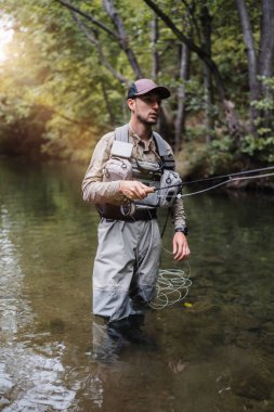 Angler dressed in fishing gear casts his line into a calm river stream surrounded by lush green forest, enjoying an outdoor fishing adventure in peaceful natural scenery