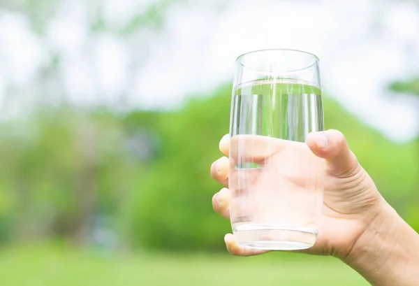Рука держит стакан чистой воды на зеленом фоне природы, здоровая концепция — стоковое фото
