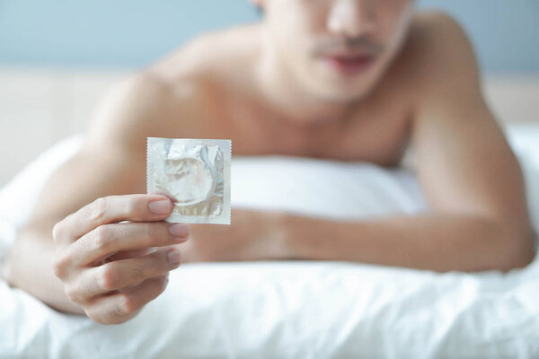 Закройте руку мужчины, держа презерватив готовым к использованию, лежа на белой кровати, здравоохранение и медицинская концепция, избирательный фокус
