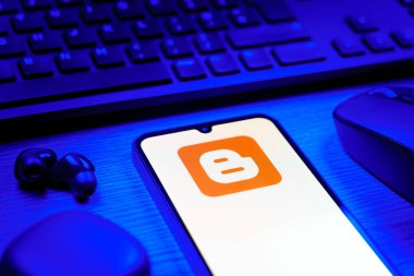 Prag, Çek Cumhuriyeti - 08 12 2025: Blogger logosunu gösteren klavyesi ve cep telefonu olan bir masanın yan görüntüsü. Blogger blog oluşturma ve yayınlama için bir platformdur.