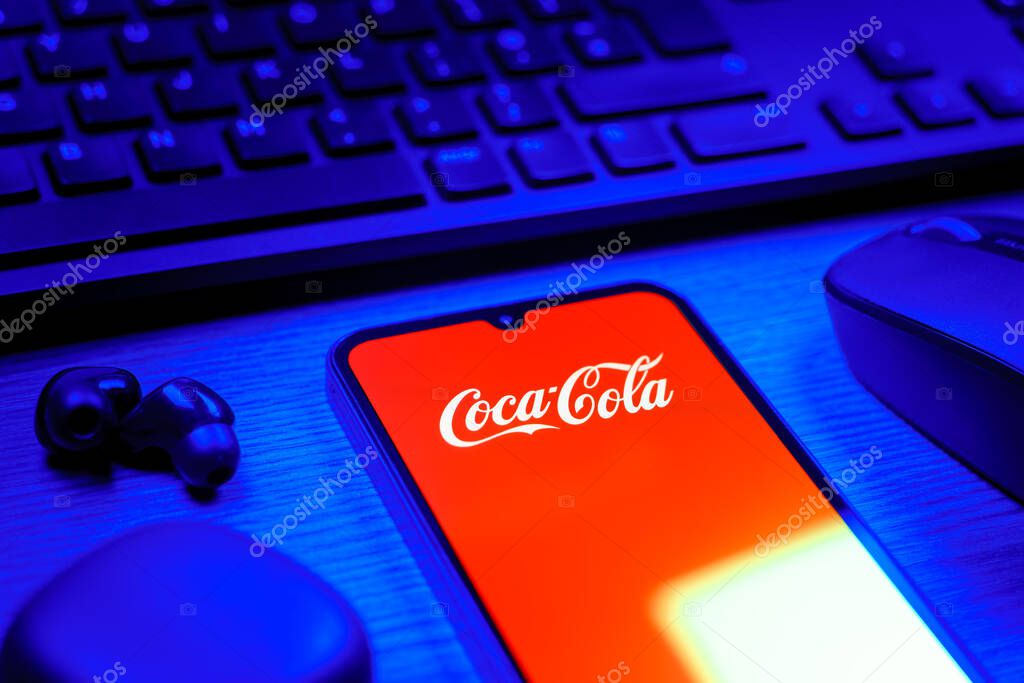 Prague, Czechia - 08 12 2025: A side view of a desk with a keyboard and a mobile phone displaying the Coca-Cola logo. Coca-Cola is a beverage company known for its soft drinks.