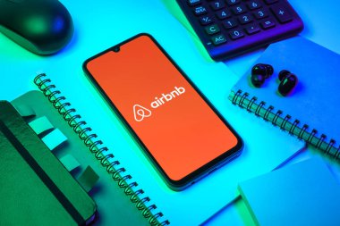 Prag, Çek Cumhuriyeti - 08 18 2025: Merkezde Airbnb logosu sergilenen bir cep telefonu ile ofis malzemeleri düzenlemesi. Airbnb dünya çapında kısa vadeli konaklama için bir platformdur..