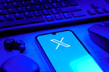 Prague, Czechia - 08 12 2025: A side view of a desk with a keyboard and a mobile phone displaying the X logo. X is a platform for sharing short posts and updates.