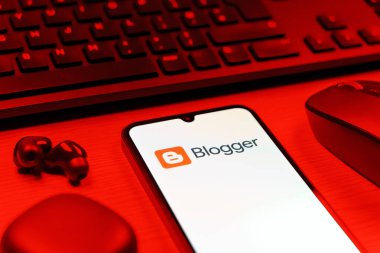Prag, Çek Cumhuriyeti - 08 12 2025: Blogger logosunu gösteren klavyesi ve cep telefonu olan bir masanın yan görüntüsü. Blogger blog oluşturma ve yayınlama için bir platformdur.