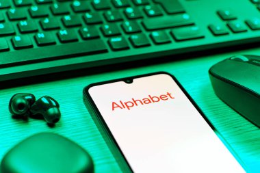 Prague, Czechia - 08 12 2025: A side view of a desk with a keyboard and a mobile phone displaying the Alphabet logo. Alphabet is a parent company of Google and other tech ventures.
