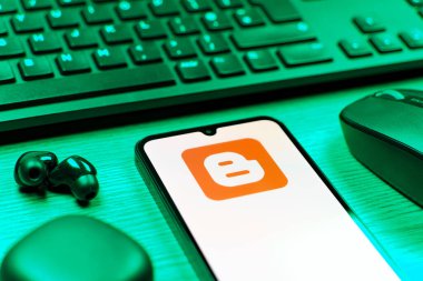 Prag, Çek Cumhuriyeti - 08 12 2025: Blogger logosunu gösteren klavyesi ve cep telefonu olan bir masanın yan görüntüsü. Blogger blog oluşturma ve yayınlama için bir platformdur.