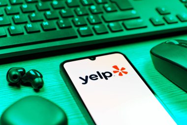 Prag, Çek Cumhuriyeti - 08 12 2025: Yelp logosunu gösteren klavyesi ve cep telefonu olan bir masanın yan görüntüsü. Yelp yerel işletmeleri bulmak ve incelemek için bir platformdur.