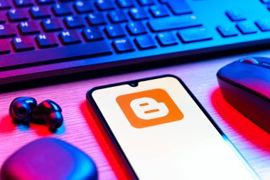 Prag, Çek Cumhuriyeti - 08 12 2025: Blogger logosunu gösteren klavyesi ve cep telefonu olan bir masanın yan görüntüsü. Blogger blog oluşturma ve yayınlama için bir platformdur.