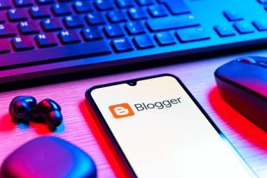 Prag, Çek Cumhuriyeti - 08 12 2025: Blogger logosunu gösteren klavyesi ve cep telefonu olan bir masanın yan görüntüsü. Blogger blog oluşturma ve yayınlama için bir platformdur.