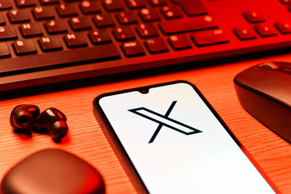 Prague, Czechia - 08 12 2025: A side view of a desk with a keyboard and a mobile phone displaying the X logo. X is a platform for sharing short posts and updates.