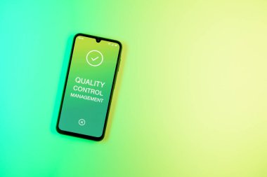 Sade arka plandaki telefon Kalite Kontrol Yönetimi metnini gösteriyor. Kalite Kontrol Yönetimi ürün veya hizmetlerin standartları karşılamasını sağlıyor.