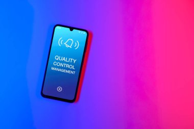 Sade arka plandaki telefon Kalite Kontrol Yönetimi metnini gösteriyor. Kalite Kontrol Yönetimi ürün veya hizmetlerin standartları karşılamasını sağlıyor.