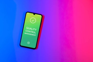 Sade arka plandaki telefon Kalite Kontrol Yönetimi metnini gösteriyor. Kalite Kontrol Yönetimi ürün veya hizmetlerin standartları karşılamasını sağlıyor.