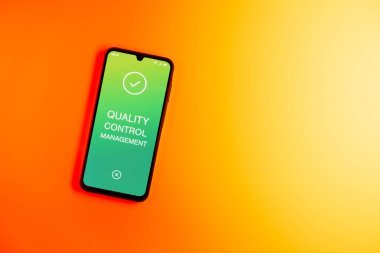 Sade arka plandaki telefon Kalite Kontrol Yönetimi metnini gösteriyor. Kalite Kontrol Yönetimi ürün veya hizmetlerin standartları karşılamasını sağlıyor.
