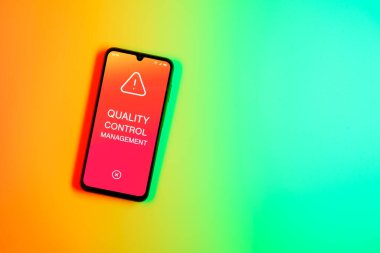 Sade arka plandaki telefon Kalite Kontrol Yönetimi metnini gösteriyor. Kalite Kontrol Yönetimi ürün veya hizmetlerin standartları karşılamasını sağlıyor.