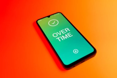Üniformalı bir arkaplanda yatan cep telefonunun fazla mesai mesajlarını gösteren bir detayı. Fazla mesai normal çalışma saatlerinin ötesinde yapılır..