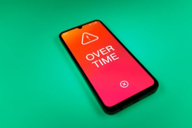 Üniformalı bir arkaplanda yatan cep telefonunun fazla mesai mesajlarını gösteren bir detayı. Fazla mesai normal çalışma saatlerinin ötesinde yapılır..