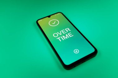 Üniformalı bir arkaplanda yatan cep telefonunun fazla mesai mesajlarını gösteren bir detayı. Fazla mesai normal çalışma saatlerinin ötesinde yapılır..