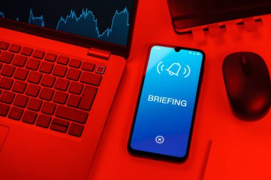 Office desk setup with laptop and phone showing Briefing text. Briefing is a short meeting or update about tasks.
