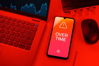 Office desk setup with laptop and phone showing Overtime text. Overtime is work done beyond regular working hours.