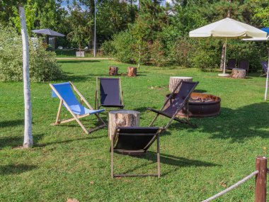 Yaz parkında şemsiyelerin altındaki yeşil çimlerin üzerinde şezlonglar, ağaç kütükleri ve ateş çukurları olan açık hava oturma alanı. Rahatlama ve rahatlama konsepti.