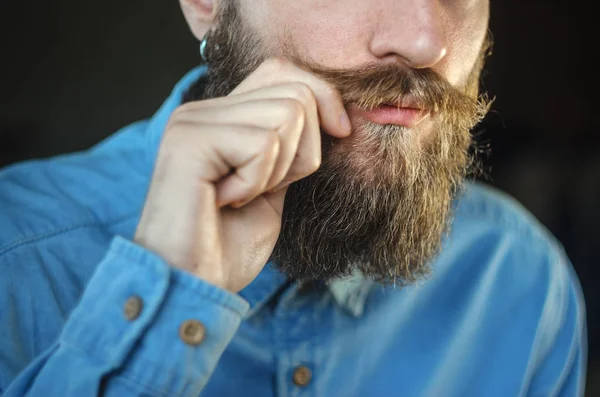 Bearded Man in a Blue Denim Shirt Twirling His Mustache with His Fingers. Portrait of a Hipster on Black Background Closeup