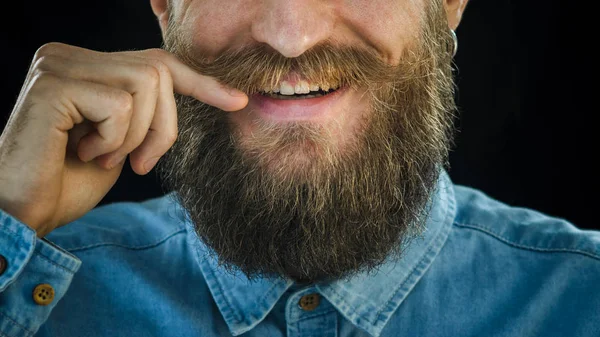 微笑着胡子的男子穿着蓝色牛仔衬衫 用手在黑色背景上扭动胡子 时髦的看 胡子和胡子特写 — 图库照片
