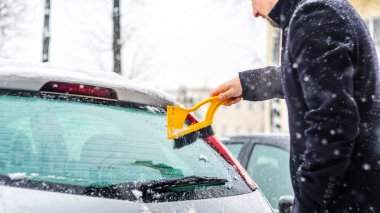 Siyah montlu genç adam arabasını sarı fırça ile kar yağışı sırasında temizler. Kış sert hava.