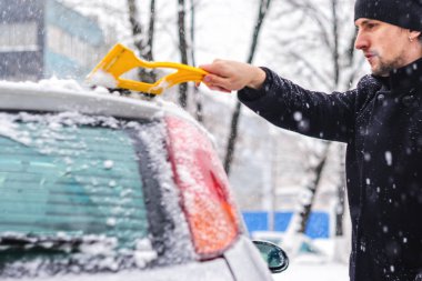 Siyah montlu genç adam arabasını sarı fırça ile kar yağışı sırasında temizler. Kış sert hava.