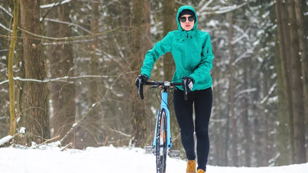 Cyclist woman goes with a bicycle on bike trails in the snowy forest in winterWinter workout outdoors concept