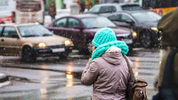 Woman in an urban environment at the crossroads talking on the phone. Huge knitted scarf and hat in turquoise color. Snowfall. Winter weather background