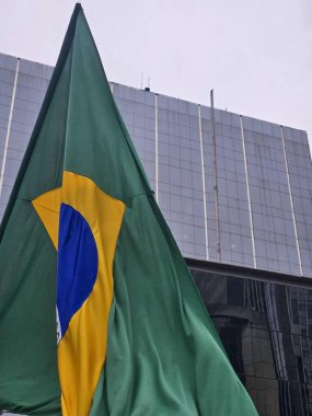 Brezilya bayrağı, parlak yeşil ve belirgin mavi ve sarı amblemi ile Paulista Bulvarı 'ndaki modern cam önlü bir binanın arka planında dalgalanır..