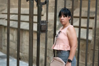 Tarihi taş duvarların ve karanlık metal kapıların önündeki kameraya bakan genç bir kadın.