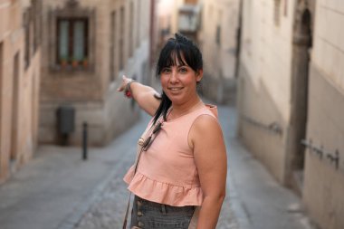 Gülümseyen kadın eli işaret ediyor, dar bir sokakta yön gösteriyor.