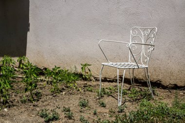 Güneşli bir bahçede, desenli bir duvarın yanında beyaz metal sandalye.