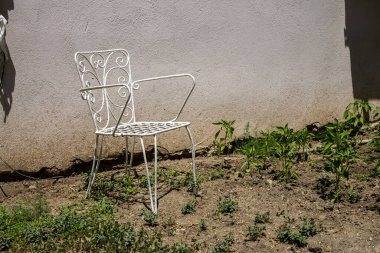 Güneşli bir bahçede, desenli bir duvarın yanında beyaz metal sandalye.