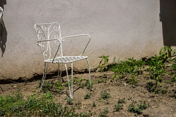Güneşli bir bahçede, desenli bir duvarın yanında beyaz metal sandalye.