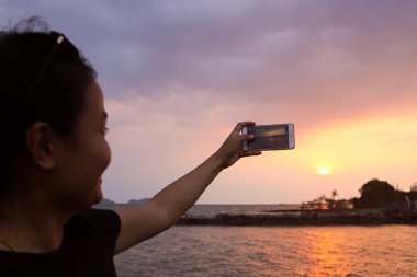 Kadın akıllı telefonuyla gün batımının fotoğrafını çekiyor.