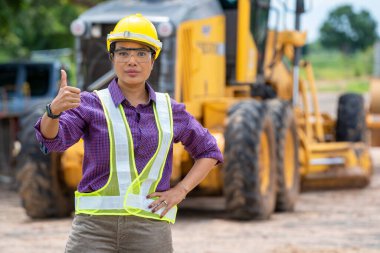 Miğfer takan kadın mühendis inşaat sahasında, inşaat sahasında, inşaat makinelerinde, buldozerlerde ve kazılarda baş parmağını gösteriyor..