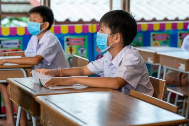 Covid 19 karantinası ve tecrit sonrasında okulda Covid 19 salgınını önlemek için hijyenik maske takan bir grup Asyalı ilkokul öğrencisi ve öğretmen..
