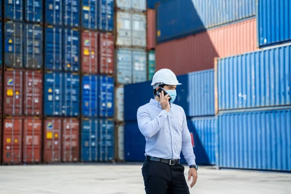 Business man wearing protective mask to Protect Against Covid-19 worker checking container box with use smart phone in shipping yard, Control loading unloading container box freight.