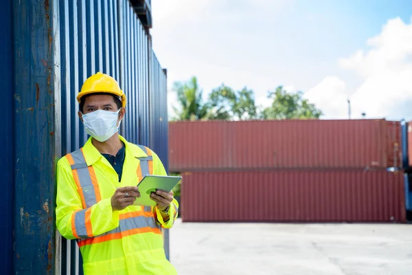 Container worker wearing protective mask to Protect Against Covid-19 working and holding digital tablet at logistic shipping cargo containers yard.