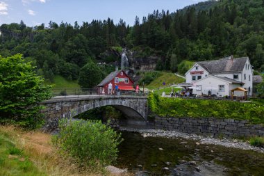 Visitors stroll across the historic stone bridge at Steinsdalsfossen in Norway, heading towards the souvenir shop and restaurant after experiencing the unique waterfall trail. The waterfall is visible in the background, cascading down the hillside.