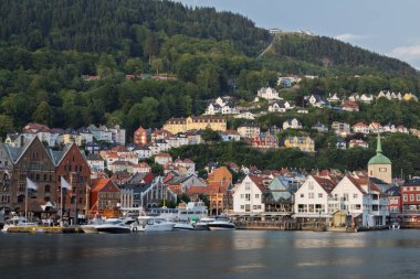 A scenic view of Bergen, Norway, showcasing the historic Bryggen wharf with its colorful wooden buildings, now housing shops and restaurants, as tourists and locals enjoy the waterfront. In the background, the lush green hillside is dotted with house