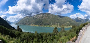 A scenic view of a long, narrow lake nestled between steep, forested mountains under a partly cloudy sky. The photographer captured this shot to showcase the natural beauty and serene atmosphere of the Norwegian landscape.